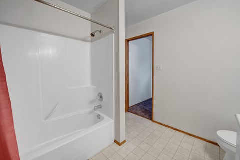 A bright bathroom tub and shower area here at Willowview Terrace with smooth white surround walls, a simple showerhead, and a sturdy curtain rod. Light tile flooring and warm wood trim lead out to the hallway, keeping the space clean, open, and easy to maintain.