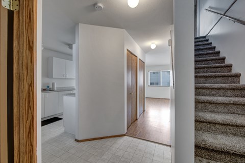A bright entry hallway here at Willowview Terrace with light tile flooring, warm wood closet doors, and a clear walkway leading into the main living area. Carpeted stairs with a simple handrail add easy access to the upper level, while the nearby kitchen cabinetry keeps the layout practical and open.