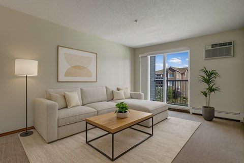 A bright living room here at Windsor Terrace featuring a light beige sectional sofa with neutral throw pillows, a modern wood coffee table, and a soft area rug for a cozy feel. A tall floor lamp, simple wall art, and a potted plant add a clean touch, while the sliding glass door brings in natural light and leads out to a private balcony view.