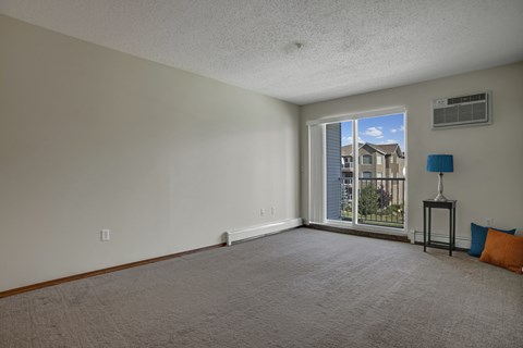 A spacious living room here at Windsor Terrace with soft carpet flooring, light neutral walls, and a large sliding glass door that opens to a private balcony with a bright outdoor view. A simple side table with a blue lamp and colorful accent pillows add a small pop of color in the corner, while a wall-mounted AC unit provides added comfort.