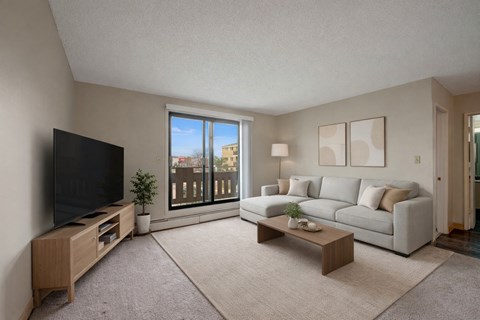 A bright living room here at Wyndham Crossing with a cozy light gray sectional sofa, neutral throw pillows, and simple wall art above the seating area. A large sliding glass door brings in natural light and opens to a private balcony, while a modern TV console, soft area rug, and small coffee table complete the clean and inviting space.