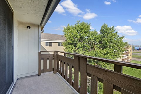 A private balcony view here at Wyndham Crossing with a concrete patio floor and a sturdy brown railing overlooking green trees and nearby buildings. The bright blue sky with soft clouds adds a fresh, open feel to the outdoor space.