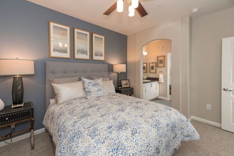A serene bedroom here at Artisan 8282 featuring a plush upholstered bed with layered neutral bedding, matching nightstands with modern lamps, soft carpeting, and a calming accent wall, complemented by ceiling fan lighting and an arched opening leading to a bright en-suite bathroom.