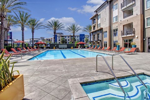 A resort-style outdoor pool here at Artisan at Main Street Metro featuring a large rectangular swimming pool with clear blue water, built-in steps and metal handrails, surrounded by red lounge chairs and umbrellas, tall palm trees, and modern apartment buildings under a bright blue sky with scattered clouds.
