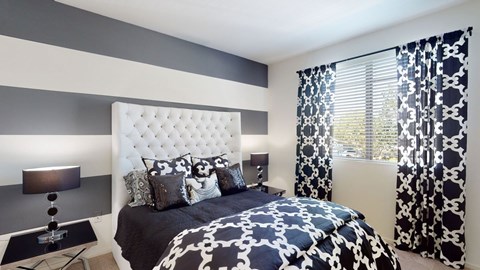 A stylish bedroom here at Artisan at Main Street Metro showcasing a tufted white headboard, bold striped accent wall in neutral tones, patterned black-and-white bedding, matching curtains framing a sunlit window, coordinated nightstands with modern lamps, and soft carpet flooring that creates a polished, inviting retreat.