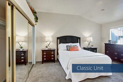 A classic bedroom here at Castlepark with plush beige carpet, neutral walls, and a neatly made bed flanked by matching nightstands and lamps. Mirrored sliding closet doors reflect light and add storage, while a window with vertical blinds brings in natural light, creating a calm and comfortable sleeping space.