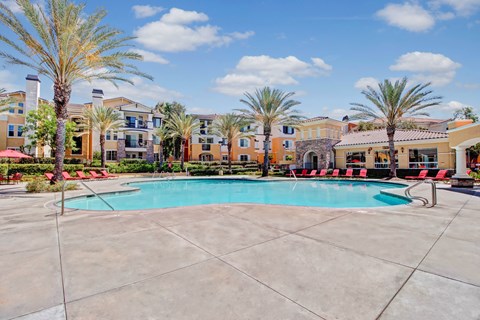 A resort-style swimming pool here at Fresco surrounded by tall palm trees, red lounge chairs, and wide concrete decking. The pool sits at the center of the community with Mediterranean-inspired apartment buildings and a clubhouse in the background, creating a bright, inviting outdoor space for relaxation and social gatherings.