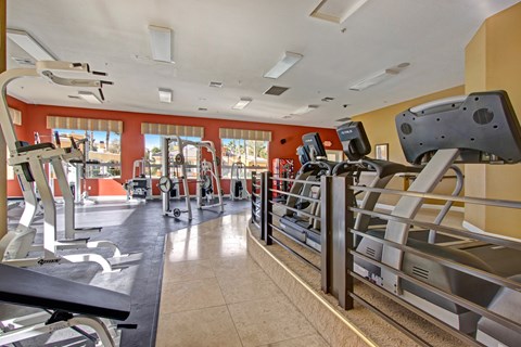A modern fitness center here at Fresco showcasing multiple cardio machines arranged along a raised platform, strength training equipment visible in the background, and large windows overlooking the community pool, complemented by warm accent walls, patterned window shades, overhead lighting, and an open layout designed for convenient daily workouts.