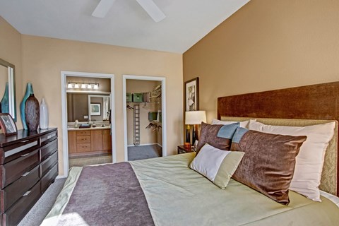 A spacious bedroom here at Fresco featuring plush carpet, a large bed with layered bedding, and a ceiling fan overhead. An open doorway leads to a walk-in closet with built-in shelving, while the ensuite bathroom with a dual-sink vanity is visible beyond, creating a comfortable and functional retreat.