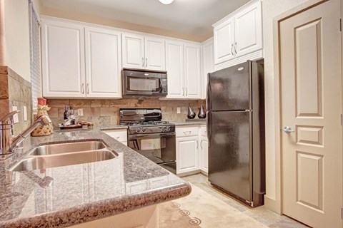 A well-appointed kitchen here at Rancho Belago featuring white shaker-style cabinetry, a stone-look backsplash, dark countertops with a double-basin sink, a built-in microwave above the range, a full-size refrigerator, and a clean, functional layout designed for everyday cooking and entertaining.