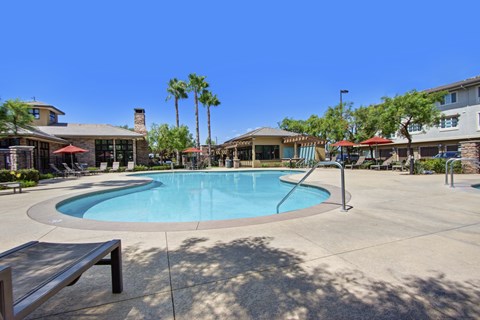 A resort-style swimming pool here at Vesada featuring a curved pool design with clear blue water, wide concrete decking, and built-in handrails for easy access. The surrounding area includes lounge chairs with umbrellas, palm trees, and shaded seating near clubhouse-style buildings, creating a relaxing outdoor space designed for leisure and warm-weather enjoyment.