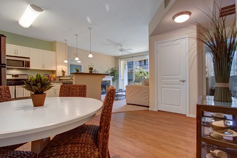 A welcoming open-concept dining and living area here at Vesada featuring warm wood-style flooring, a round dining table with woven chairs, and a bright kitchen with white cabinetry and pendant lighting. The space flows into a comfortable living room with a fireplace and sliding glass doors that open to a private balcony, filling the home with natural light.