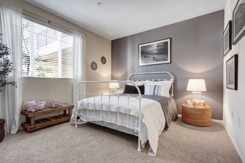 A serene bedroom here at Vesada showcasing a white metal bed frame dressed in crisp white bedding with soft gray and neutral accent pillows, set against a calming gray accent wall. The room features plush carpeting, matching bedside lamps on woven and wood tables, framed wall art, and a large window with sheer curtains that fills the space with natural light.