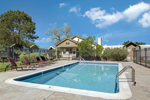 A bright outdoor pool here at Austin Park featuring a sparkling rectangular swimming pool with clear blue water, surrounding concrete deck with metal handrails, lounge chairs lined along the fence, and landscaped greenery, creating a refreshing community space under a sunny blue sky.