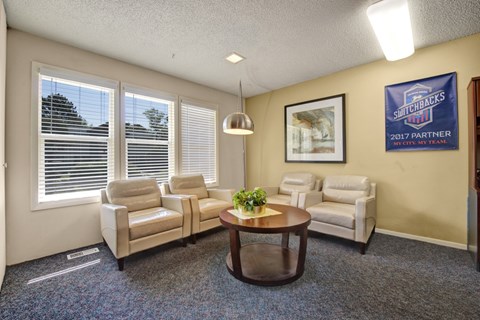 A cozy community lounge living room here at Austin Park featuring three cream leather armchairs arranged around a round wooden coffee table with greenery, soft carpet flooring, large windows with white blinds bringing in natural light, and warm accent walls with framed artwork and decor.