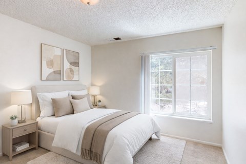 A bright, airy bedroom here at Austin Park featuring a softly upholstered bed with crisp white bedding, neutral accent pillows, matching bedside tables with modern lamps, abstract wall art above the headboard, plush carpet flooring, and a large window with vertical blinds that fills the space with natural light.