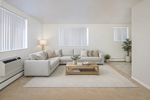 A bright modern living room here at Bristol Square featuring a spacious light gray sectional sofa with neutral throw pillows, a wooden coffee table on a soft area rug, vertical blinds allowing natural light, light-toned walls, and clean finishes that create an open, inviting atmosphere for everyday living and relaxing.