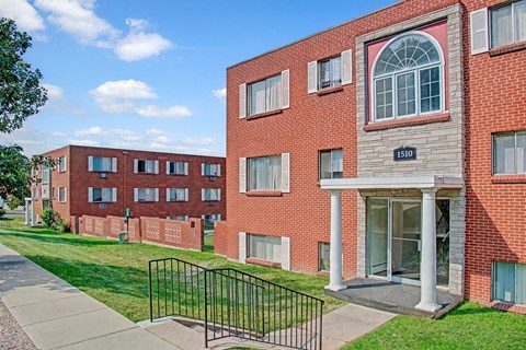 A classic brick apartment building exterior here at Bristol Square featuring a main entrance with white columns, an arched window above the doorway, multiple large windows with shutters, well-kept green lawns, a concrete walkway with railings, and additional residential buildings visible in the background under a bright blue sky.