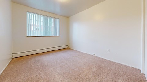 A bright, unfurnished bedroom here at Bristol Square featuring neutral carpet flooring, soft white walls, and a wide window with blinds that brings in natural light, creating a clean, open space ready for personal touches.