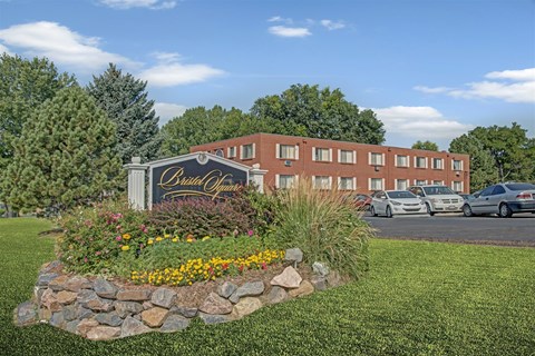 A welcoming apartment community entrance sign here at Bristol Square showcasing a landscaped monument sign surrounded by colorful flowers, decorative stone edging, green lawn, mature trees, parked vehicles, and classic brick apartment buildings beneath a bright blue sky.