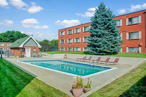 A sunny outdoor swimming pool here at Bristol Square featuring a rectangular pool surrounded by a concrete deck, red lounge chairs, green lawn landscaping, and a tall evergreen tree set against brick apartment buildings and a bright blue sky with scattered clouds.