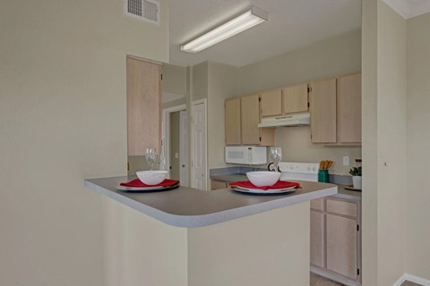 A bright kitchen here at Camelback Pointe featuring light wood cabinetry, neutral countertops, and a raised breakfast bar set with place settings, with a white microwave and stove beneath a vent hood, overhead lighting, and an open layout that connects easily to the rest of the apartment.