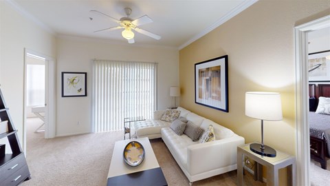 A bright, furnished living room here at Camelback Pointe featuring a white sofa, a ceiling fan, framed artwork, a table lamp, and a wide window with vertical blinds that fills the space with natural light.