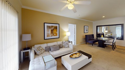 A spacious living and dining area here at Camelback Pointe featuring a white sectional sofa, a modern coffee table, a ceiling fan, framed artwork, and a dining table set near a large wall mirror.