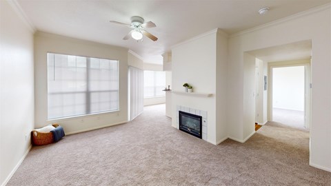 A bright, open living room here at Camelback Pointe featuring neutral carpet, a corner fireplace, a ceiling fan, and large windows with blinds that bring in natural light.