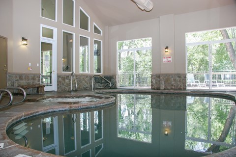 A serene indoor pool here at Cheyenne Creek Luxury featuring a curved pool edge with stone detailing, clear reflective water, tall windows that bring in abundant natural light and views of surrounding trees, stone accent walls, metal handrails for easy entry, and a calm, spa-like atmosphere designed for year-round relaxation.