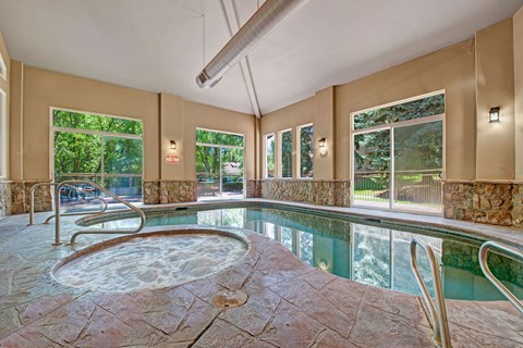 A bright indoor pool here at Cheyenne Creek Luxury featuring a connected spa with bubbling water, stone-accent pool deck, metal handrails for easy entry, large windows that fill the space with natural light, and peaceful views of surrounding trees creating a relaxing, resort-style atmosphere.