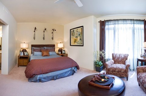 A cozy bedroom here at Cheyenne Creek Luxury showcasing a comfortable bed with layered bedding, matching nightstands with lamps, plush carpeting, and soft neutral walls, complemented by a sitting area with upholstered chairs, natural light from a large window with curtains, and tasteful wall art creating a relaxing retreat.