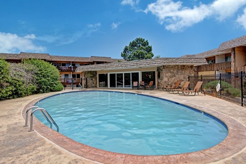 A refreshing outdoor pool here at Copper Stone featuring a curved swimming pool with clear blue water, surrounding concrete deck space, lounge chairs for relaxing, and a stone-accented pool house, all set within a landscaped courtyard under open skies.