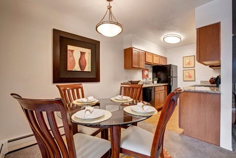 A welcoming dining area here at Copper Stone featuring a round glass-top table set for four with wooden chairs and upholstered seats, positioned beside an open kitchen with warm wood cabinetry, sleek countertops, modern appliances, pendant lighting, and framed wall art that creates a polished and comfortable apartment dining space.