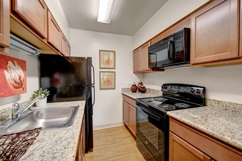 A bright galley-style kitchen here at Copper Stone featuring warm wood cabinetry, speckled countertops, a stainless sink, and black appliances arranged for efficient cooking, with wood-look flooring, overhead lighting, simple wall art, and clean lines that create a functional and inviting apartment kitchen space.