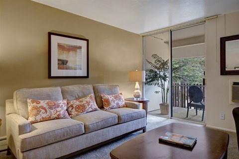 A cozy living room here at Copper Stone featuring a neutral fabric sofa with patterned accent pillows, a wooden coffee table, and framed wall art, complemented by soft carpeting, a table lamp for warm lighting, and a sliding glass door that opens to a private balcony overlooking mature greenery.