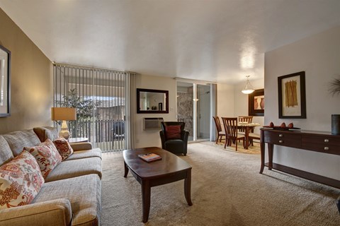 A spacious open living room here at Copper Stone showcasing a neutral sofa with patterned accent pillows, plush carpeting, a wooden coffee table, and a cozy armchair, flowing into an adjacent dining area with a wood dining table and chairs, accented by framed artwork, warm lighting, and sliding glass doors that bring in natural light.