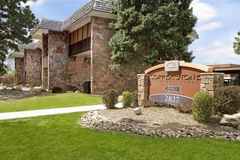 A welcoming exterior monument sign here at Copper Stone featuring a stone base and curved signage set along a manicured lawn, with mature evergreen trees, neatly trimmed shrubs, and stone-accented apartment buildings creating a polished and inviting community entrance.