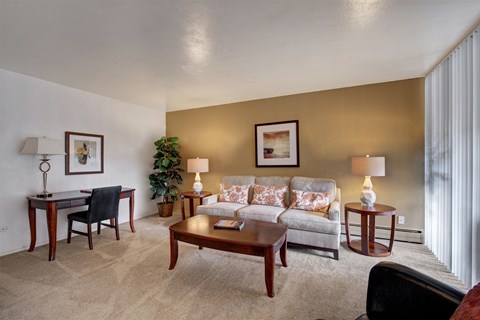 A spacious living room here at Copper Stone showcasing a neutral sofa with patterned accent pillows, a wooden coffee table, plush carpeting, and coordinated accent furniture, opening into a dining area and kitchen, with warm lighting, framed artwork, and a small workspace creating a comfortable and inviting apartment layout.