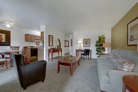 A spacious living room here at Copper Stone showcasing a neutral sofa with patterned accent pillows, a wooden coffee table, plush carpeting, and coordinated accent furniture, opening into a dining area and kitchen, with warm lighting, framed artwork, and a small workspace creating a comfortable and inviting apartment layout.