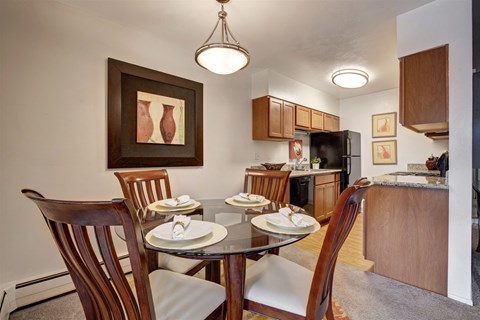 A welcoming dining area here at Copper Stone featuring a round glass-top table set for four with wooden chairs and upholstered seats, positioned beside an open kitchen with warm wood cabinetry, sleek countertops, modern appliances, pendant lighting, and framed wall art that creates a polished and comfortable apartment dining space.