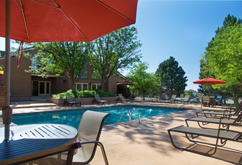 A bright outdoor pool area here at Eagle Ridge featuring a rectangular swimming pool with clear blue water, surrounding concrete deck, multiple lounge chairs arranged along the poolside, patio tables with red umbrellas, mature trees providing shade, and a clubhouse building visible in the background under a sunny blue sky.