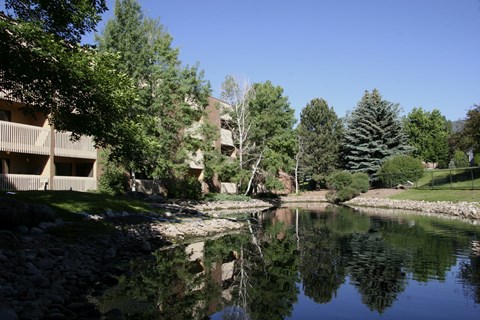 A peaceful landscaped pond here at Eagle Ridge reflecting surrounding trees and blue sky, with gently sloped grassy banks, natural stone edging, and mature evergreens alongside a brick apartment building with balconies, creating a serene outdoor setting that highlights the community’s greenery and scenic water feature.