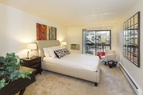 A bright bedroom here at Eagle Ridge featuring a spacious layout with a modern upholstered bed dressed in neutral bedding, a dark wood nightstand with a table lamp, soft carpeted flooring, and a cozy accent chair near a sliding glass door that opens to a private balcony, allowing natural light to fill the room and create a calm, comfortable retreat.