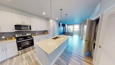 A modern open kitchen here at Experience at Epicenter featuring white shaker-style cabinets, sleek countertops, and a large center island with an integrated sink and pendant lighting. Stainless-steel appliances line the kitchen wall, while wood-style flooring flows into the open living area. An in-unit laundry closet and balcony access add everyday convenience.