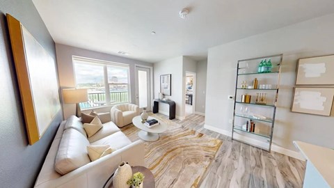 A contemporary living room here at Experience at Epicenter showcasing wood-style flooring, a light neutral sofa with accent pillows, a matching armchair, a round coffee table styled with decor, an abstract area rug, a floor lamp, framed wall art, open shelving with decorative accents, and large windows with a glass door that fill the space with natural light and offer balcony views.