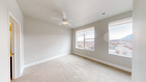 A spacious bedroom here at Experience at Epicenter featuring plush carpet, soft neutral walls, and a ceiling fan centered overhead. Two large windows with simple shades bring in natural light and showcase city and mountain views, while the open layout provides flexible space for a bed, nightstands, and additional furniture.