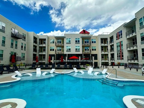 A resort-style outdoor pool here at Experience at Epicenter featuring a spacious swimming area with in-water lounge chairs, surrounding poolside seating, and red umbrellas for shade. Modern apartment buildings with balconies frame the courtyard, creating a vibrant community setting ideal for relaxing, socializing, and enjoying sunny days.