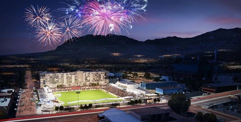 A celebratory aerial scene here at Experience at Epicenter featuring a brightly lit sports field filled with spectators, surrounded by modern apartment buildings and bustling streets. Colorful fireworks burst above the nearby mountains against a twilight sky, capturing the energy, entertainment, and vibrant community atmosphere of the neighborhood.