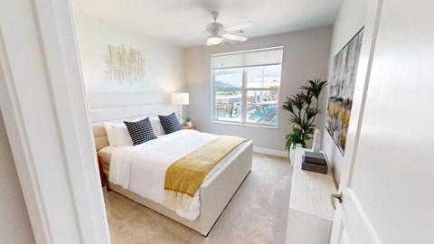 A bright bedroom here at Experience at Epicenter featuring plush carpet, soft neutral walls, and an upholstered bed styled with crisp white bedding and textured accent pillows. A ceiling fan hangs overhead, while a large window with simple shades offers city and mountain views. A sleek dresser, modern wall art, and greenery complete the inviting space.
